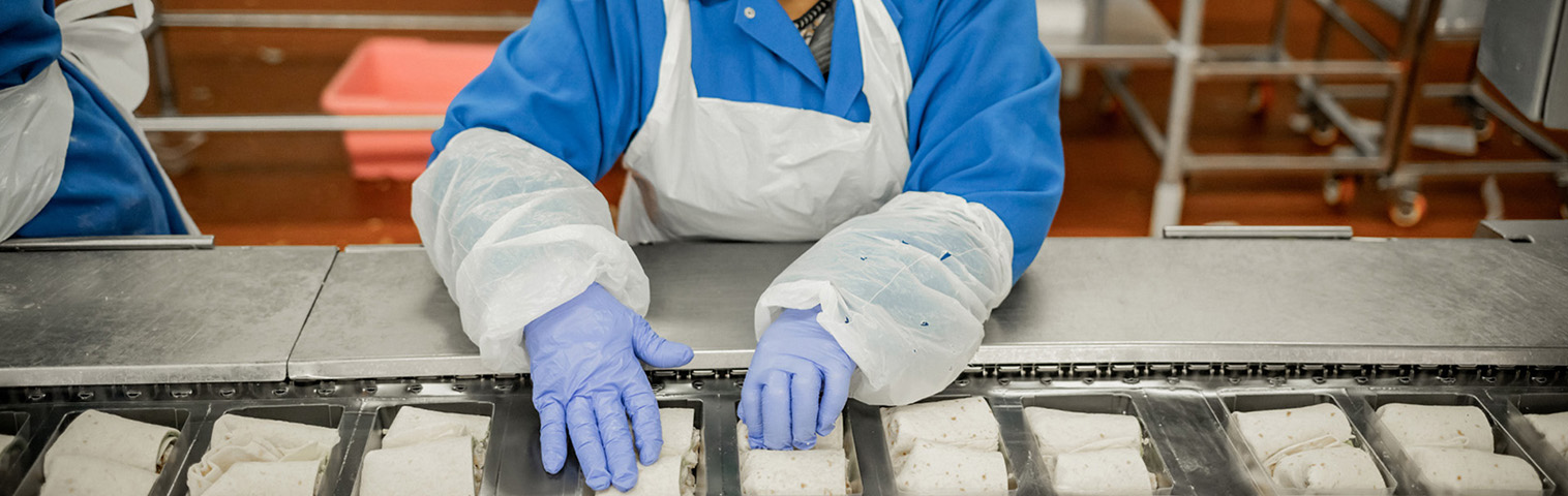 A top down view of an employee wearing an apron, gloves, hairnet and mask securing wraps in trays on an assembly line