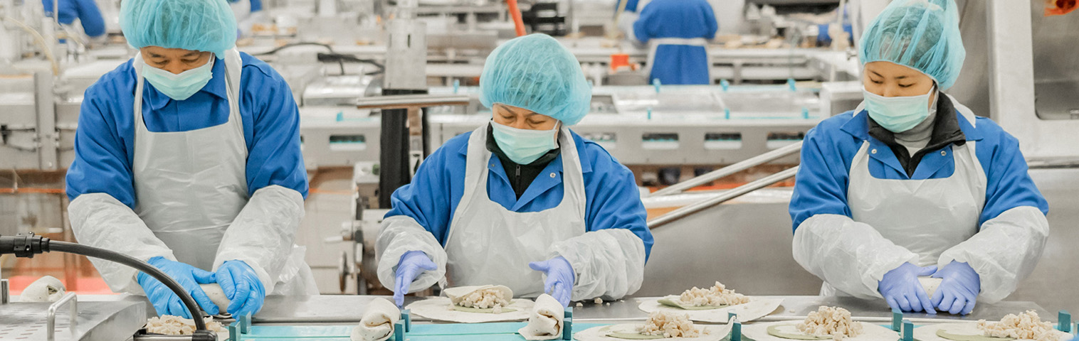 A front on view of three employees in aprons, gloves, hairnets and masks assembling chicken salad wraps